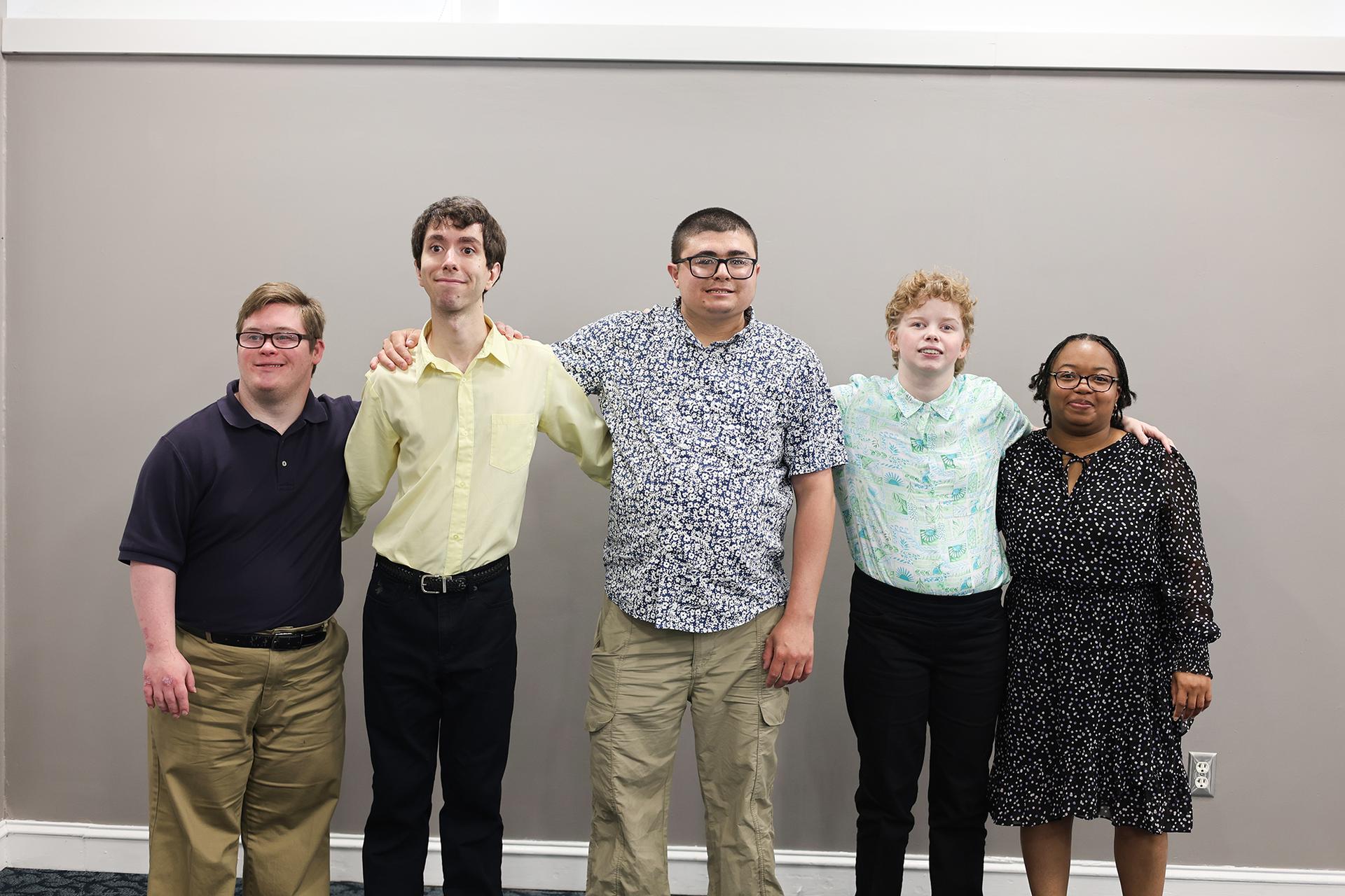 A group of people stand in front of a gray wall with their arms around each other.
