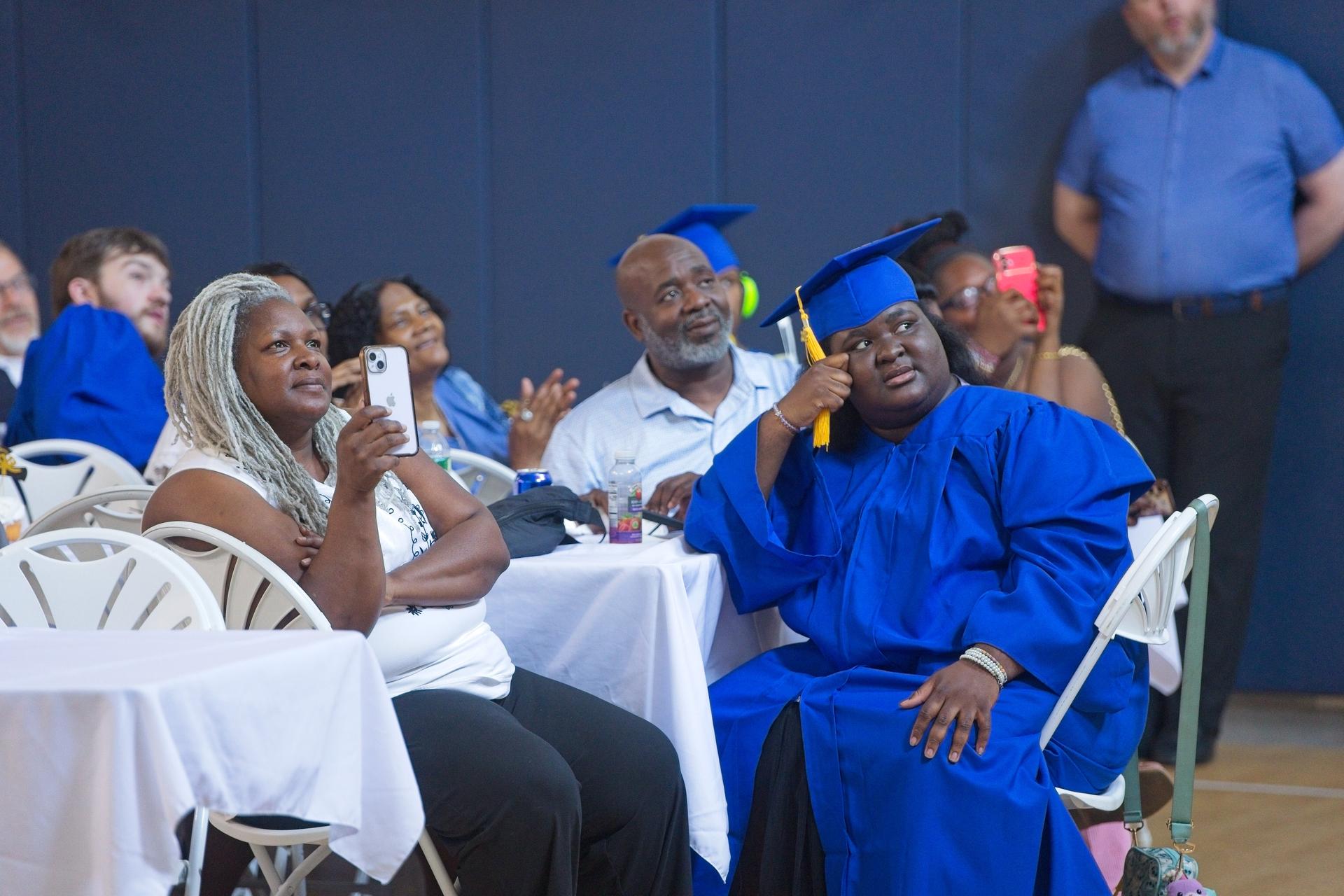 A young female graduate in blue robes sits at a table with her family during a graduation ceremony.