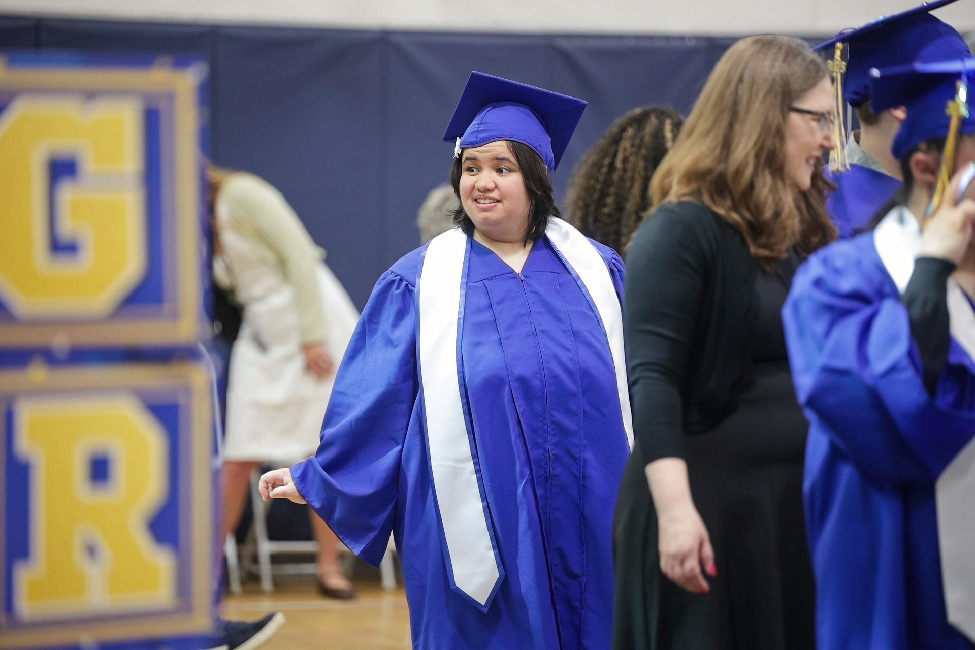 A young woman wears a blue graduation cap and gown with a white sash.