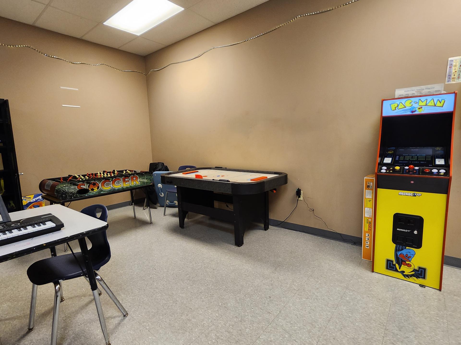 A Ms. Pac-Man machine, air hockey, foosball, and a table with a small keyboard on it in a cream colored room.
