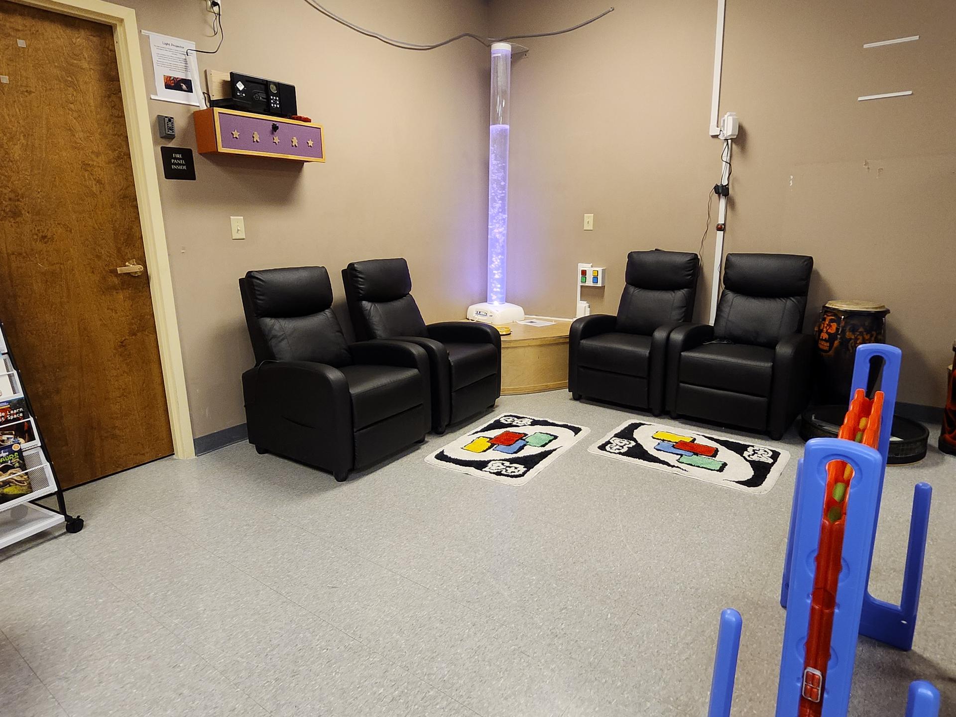 Four black massage chairs with a large Connect 4 game, and sensory lighting in a cream colored room.
