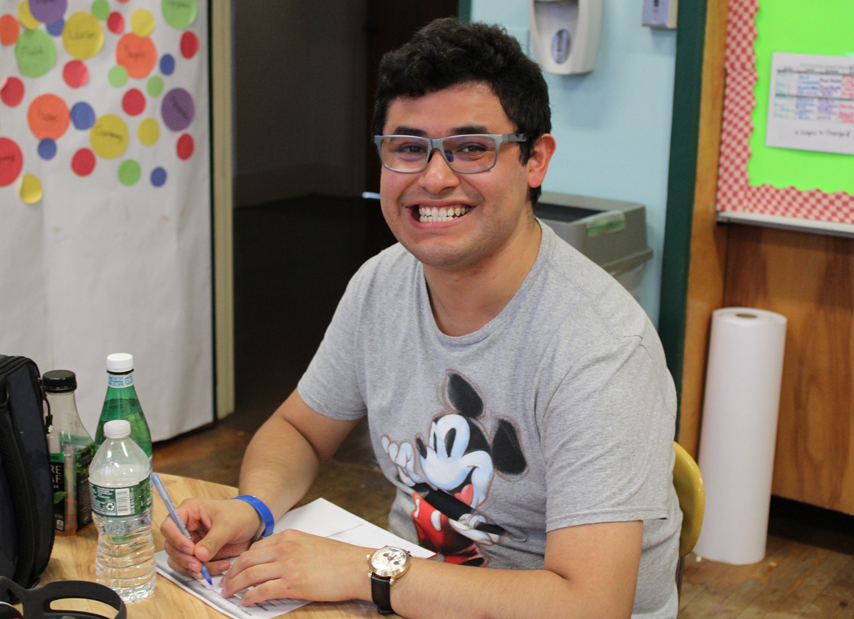 A young man with glasses wearing a gray t-shirt with Mickey Mouse on it smiles as he sits at a table.