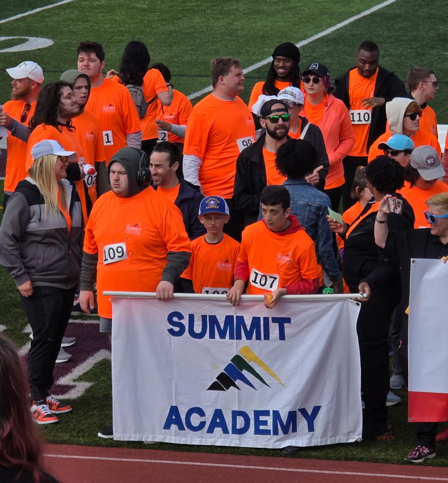 A group of people in orange t-shirts gather behind a banner with the words Summit Academy on it.