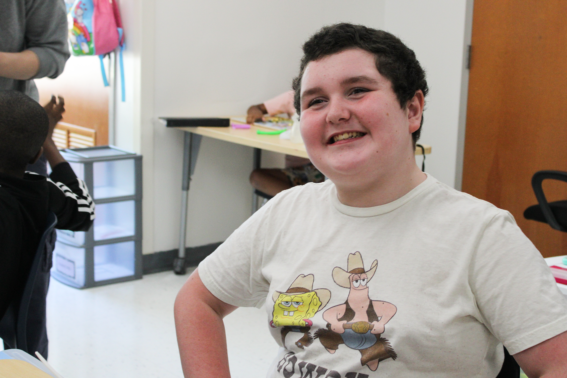 A boy wearing a white shirt smiles in a classroom.