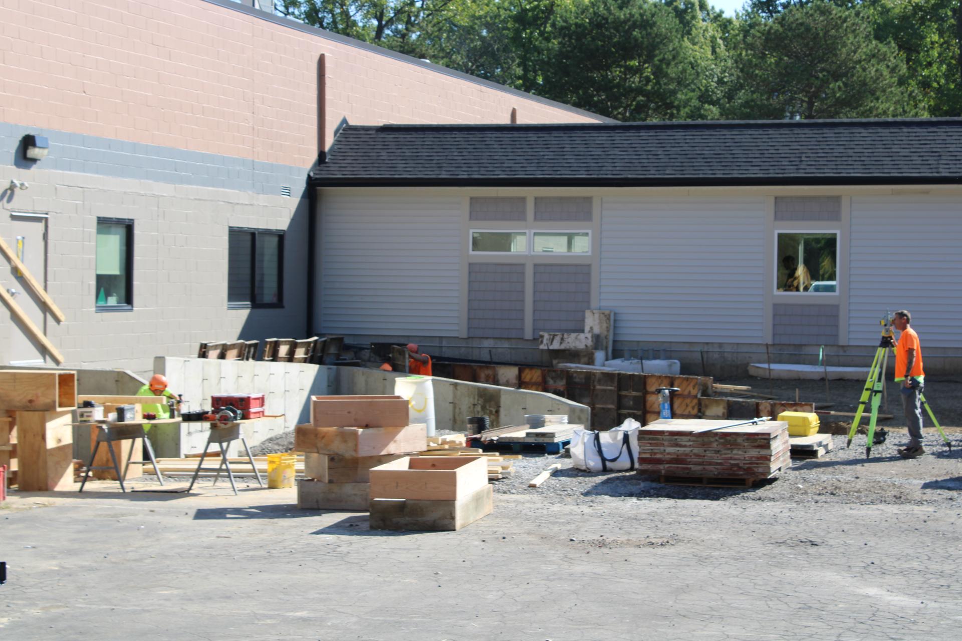 Construction materials are piled around a hole outside a white building.