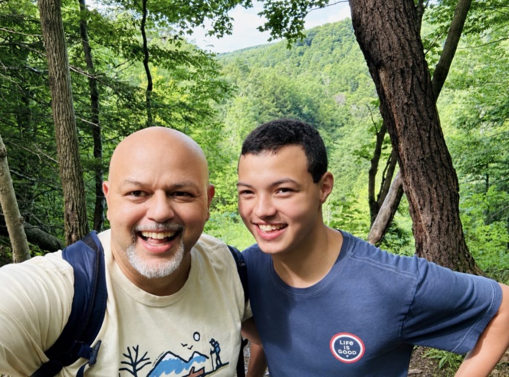 A man and his son pose on a hiking trail surrounded by trees.