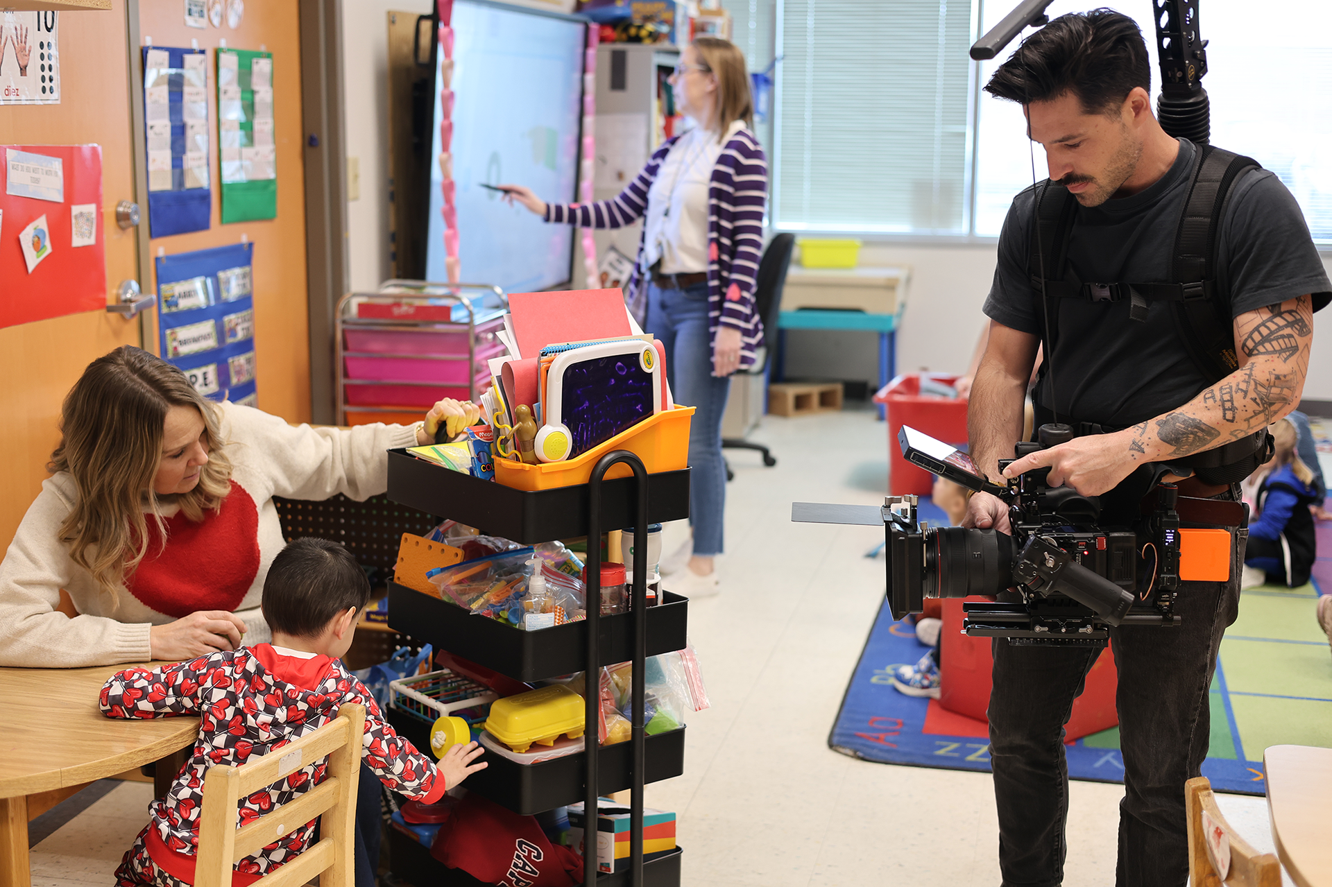 A man with a camera films a student and teacher at a table in a preschool classroom.