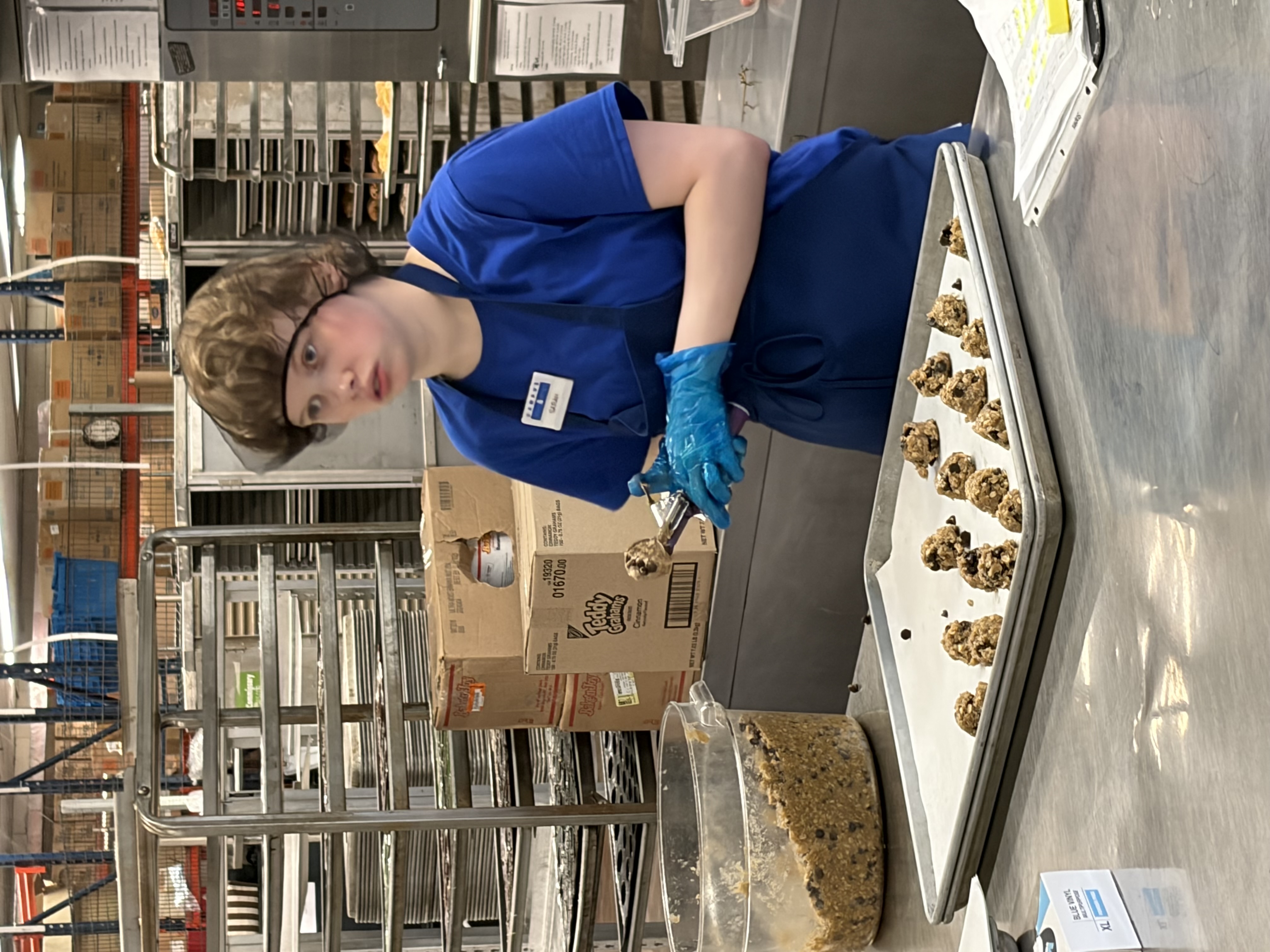 A young woman wearing a hairnet and blue shirt and apron scoops cookie dough onto a baking sheet in a kitchen.