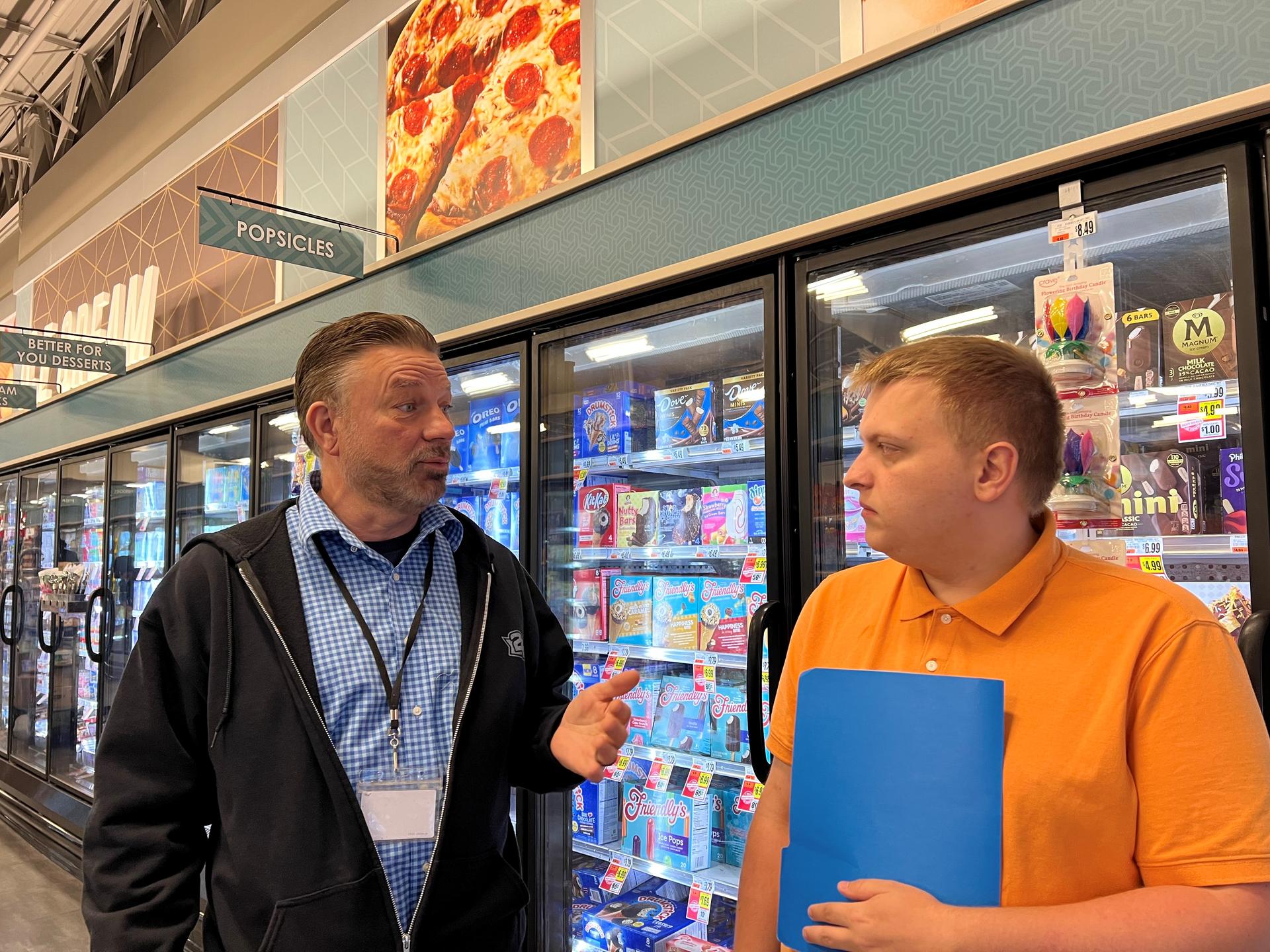 Two men dressed in work attire talk in the aisle of a grocery store.