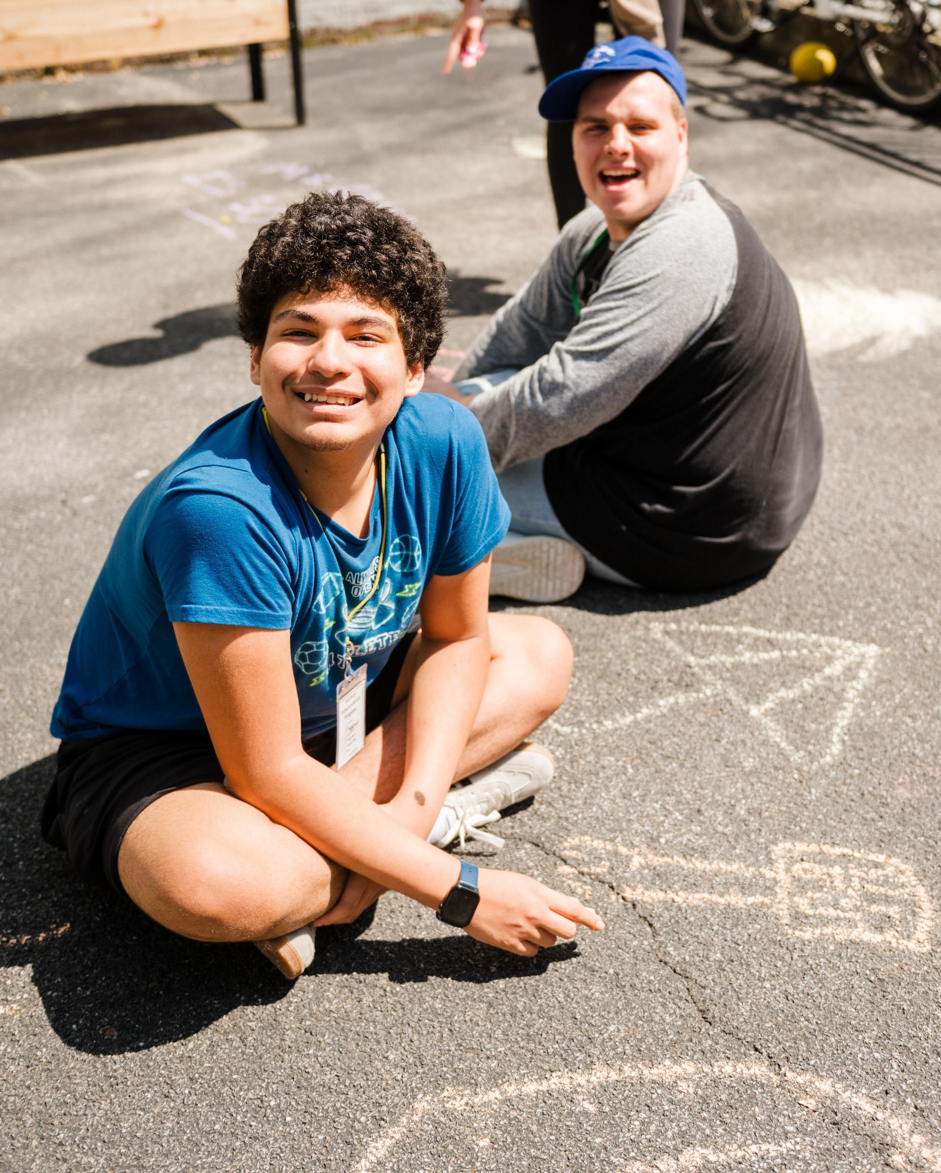 Two boys draw with chalk on the pavement outside.