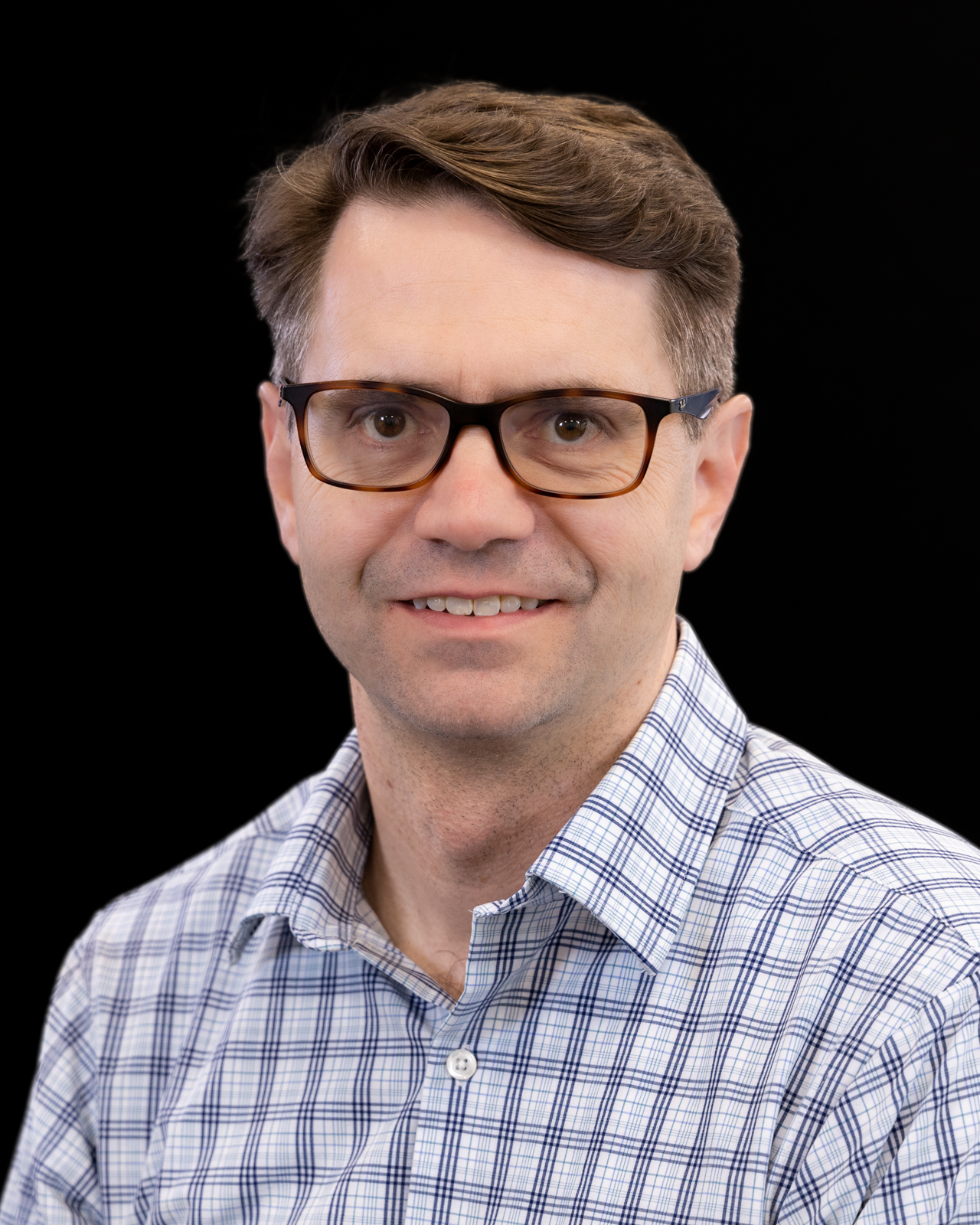 A man with glasses wearing a blue and white checked shirt in front of a black background
