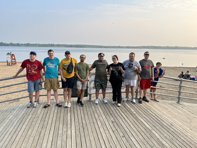 A group of people pose on a boardwalk with sand and water behind them.