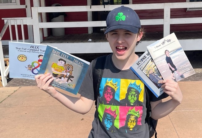 A young man wearing a black t-shirt and baseball cap holds up four children's books.
