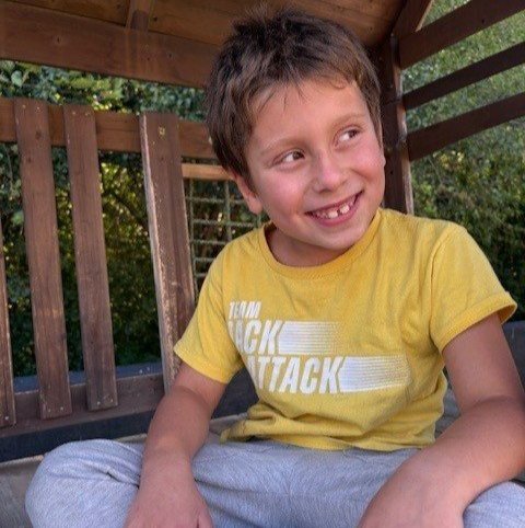 A young boy wearing a yellow t-shirt and gray sweatpants sits in a treehouse.