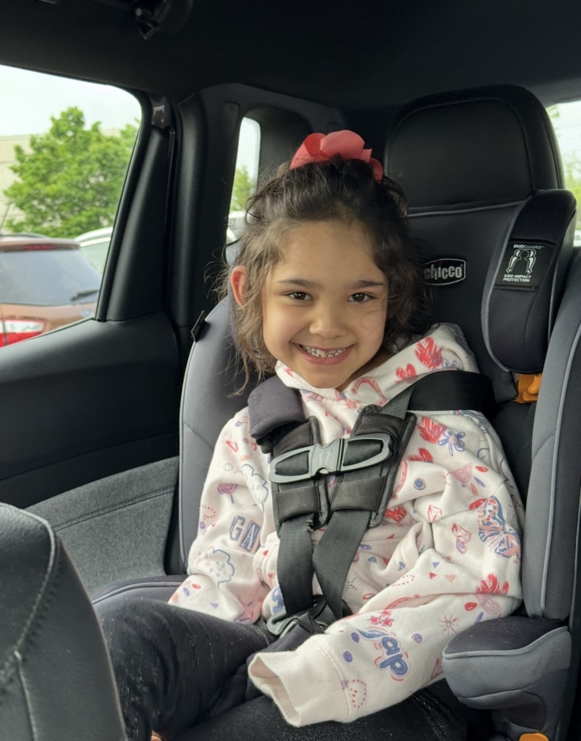A young girl wearing a white patterned sweatshirt in a car seat smiles.