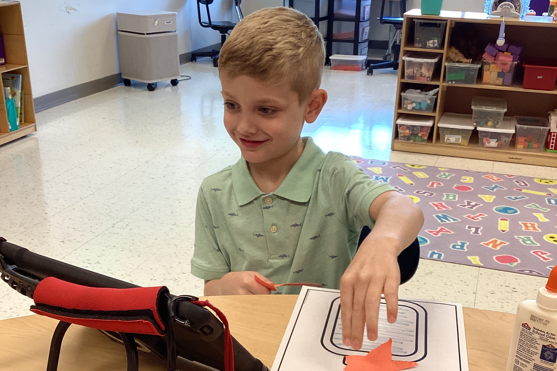 A boy smiles as he works on a craft and looks at his iPad at a desk.