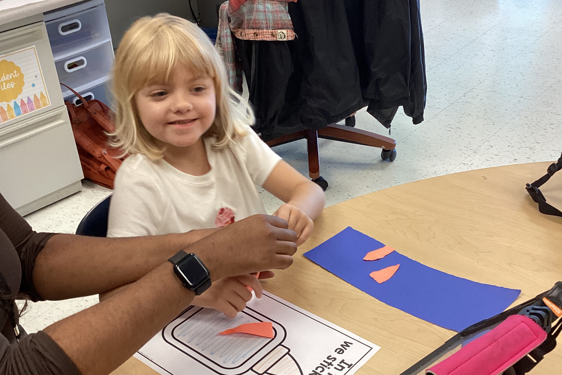 A girl smiles as she works on a craft at a school desk.
