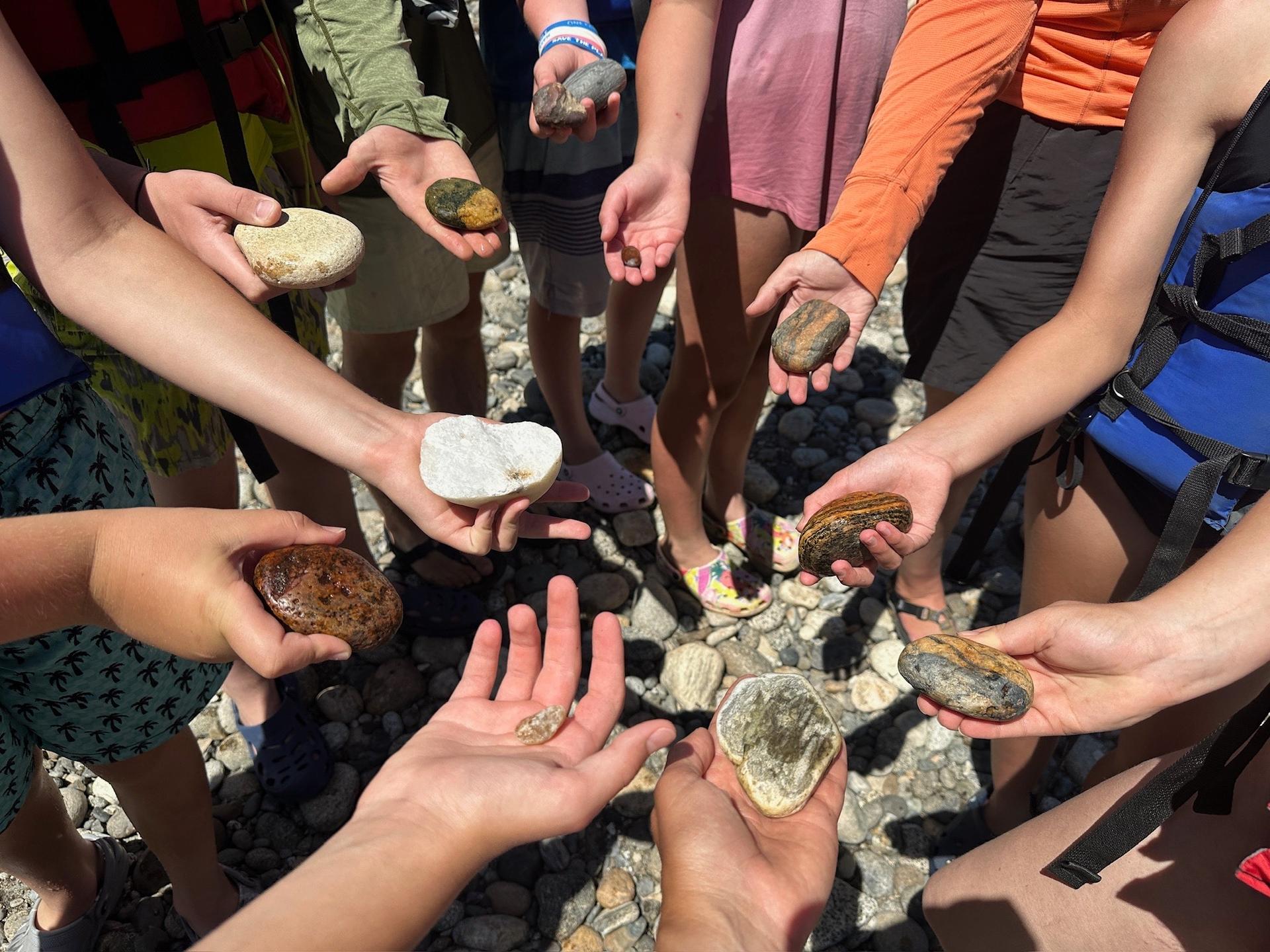 A picture of summer campers standing in a circle on a river bank holder rocks in their palms.
