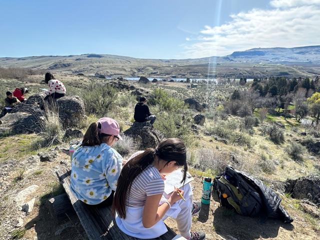 Students sitting on a bench overlooking a landscape with clip boards.
