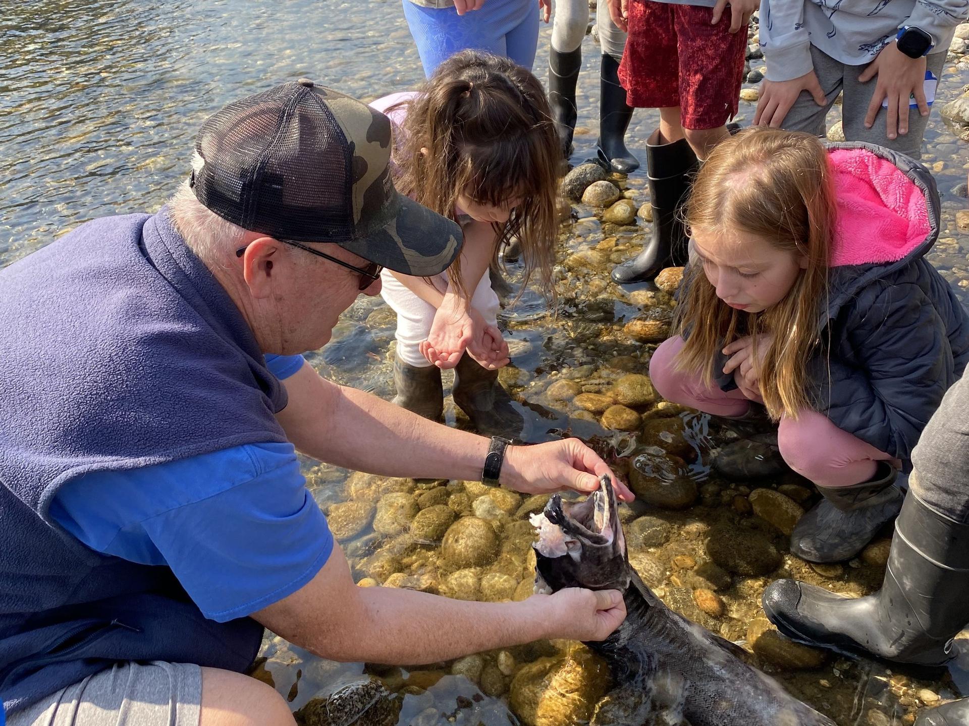 A group gathers around a volunteer near the river as he holds a dead salmon for students to view.