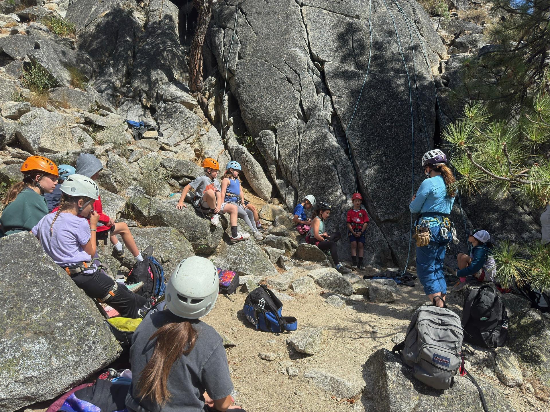 WRI Rock On! Camp photo of campers and instructors sitting below an outdoor rock wall with helmets and ropes set up waiting to climb.