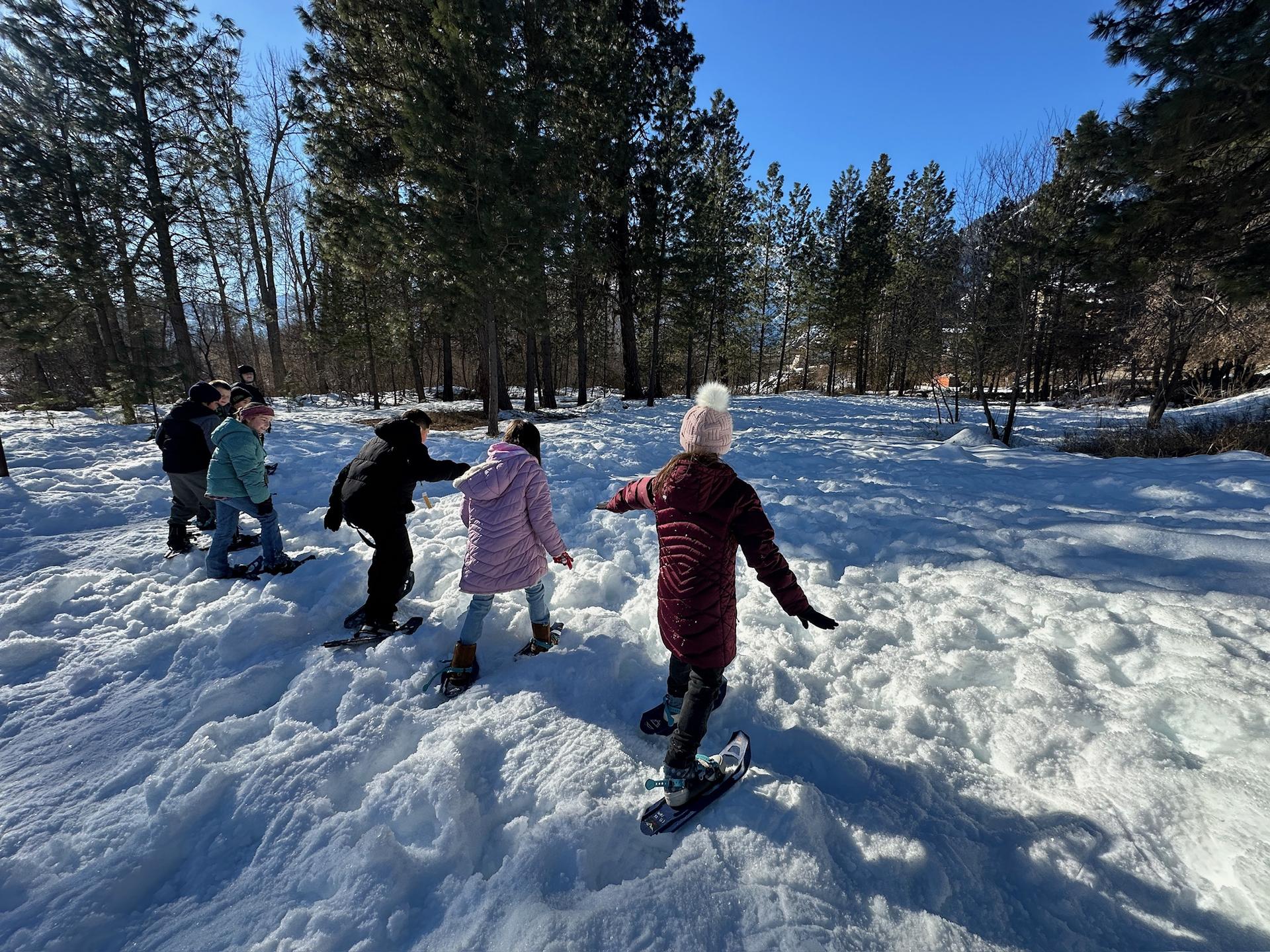 Kids running in a line with snowshoes through an open, snowy field.