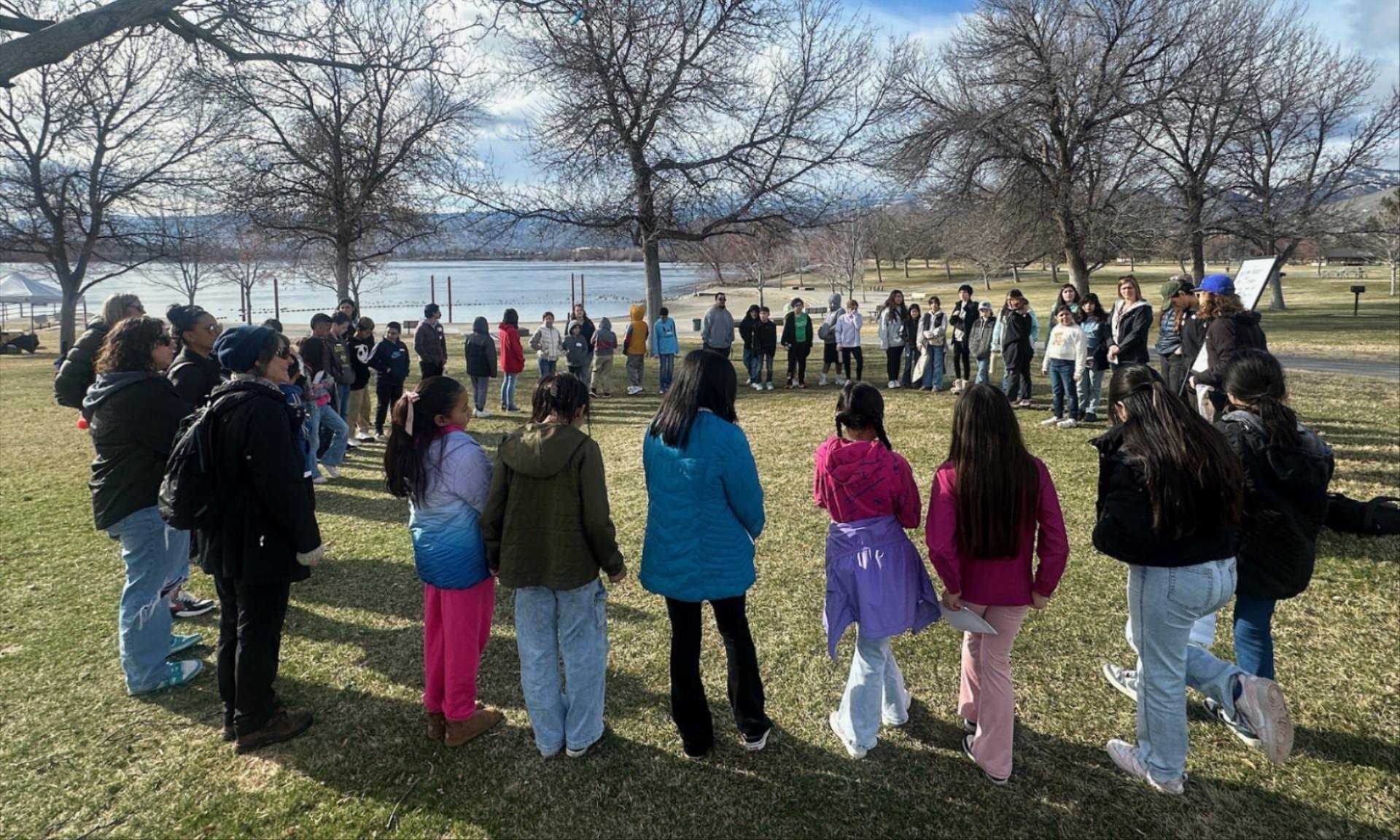 Picture from Wenatchee River Institute's Traveling Naturalist program. A group of students standing in a circle outside in the fall in an outdoor park near a river.