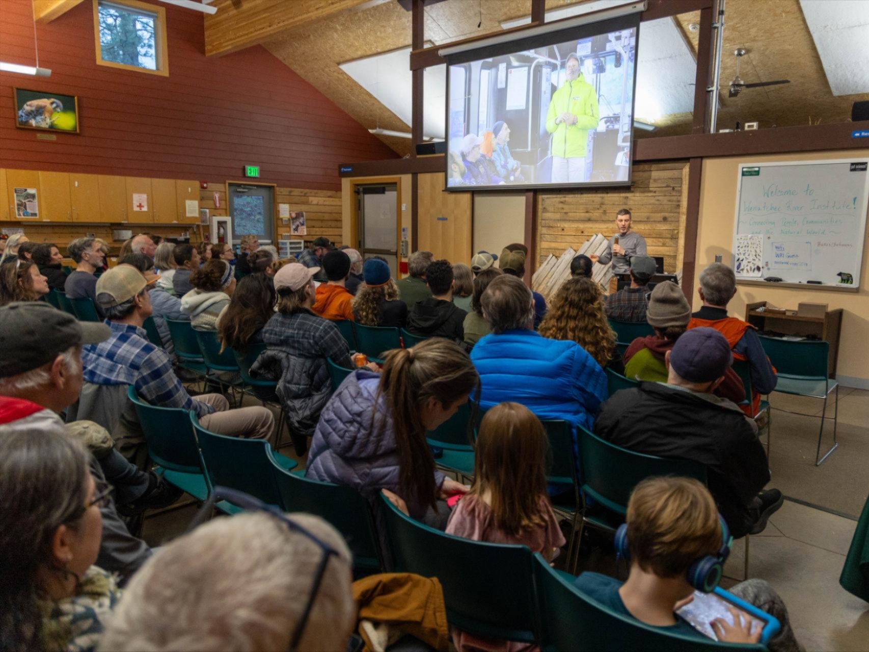 A group of community members gathered in the Red Barn looking towards the speaker and screen.